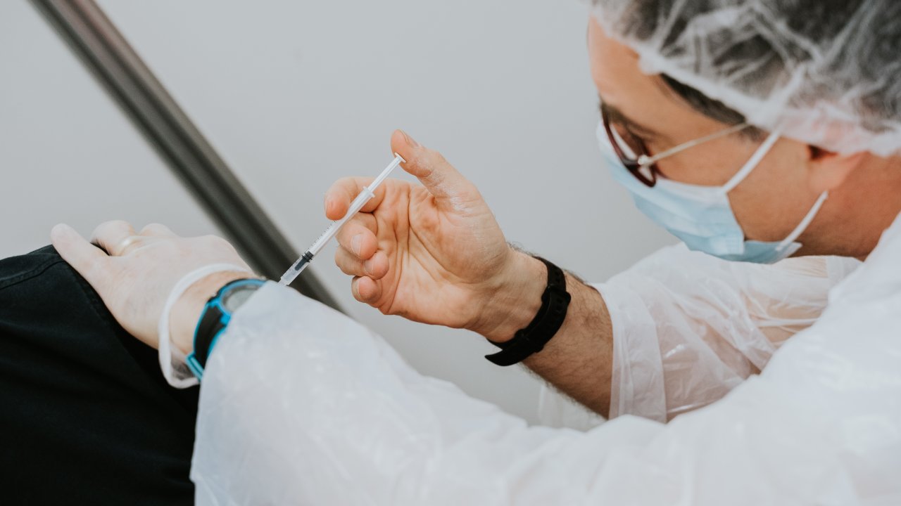 Foto de homem com equipamentos de proteção aplicando vacina da gripe em braço.